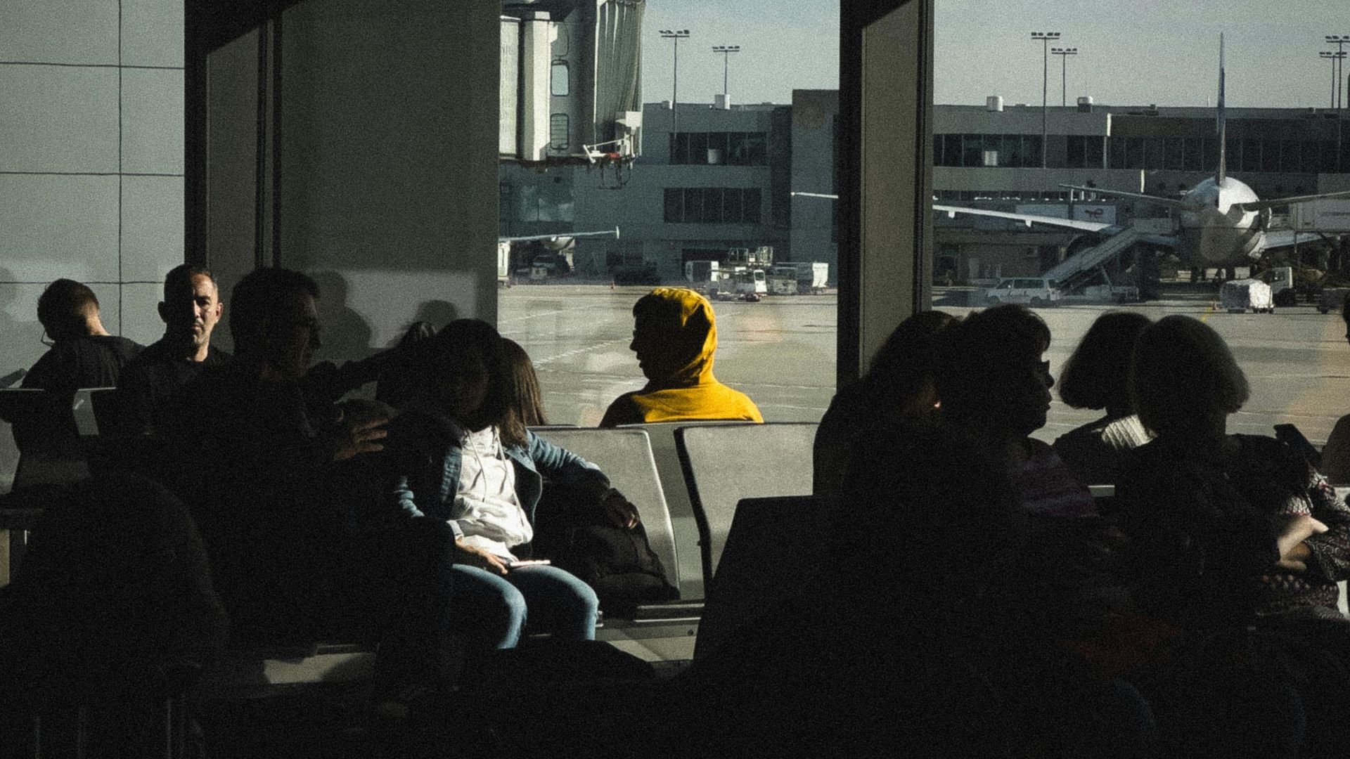 Silhouetted travelers in an airport terminal waiting area near large windows with outside tarmac view.