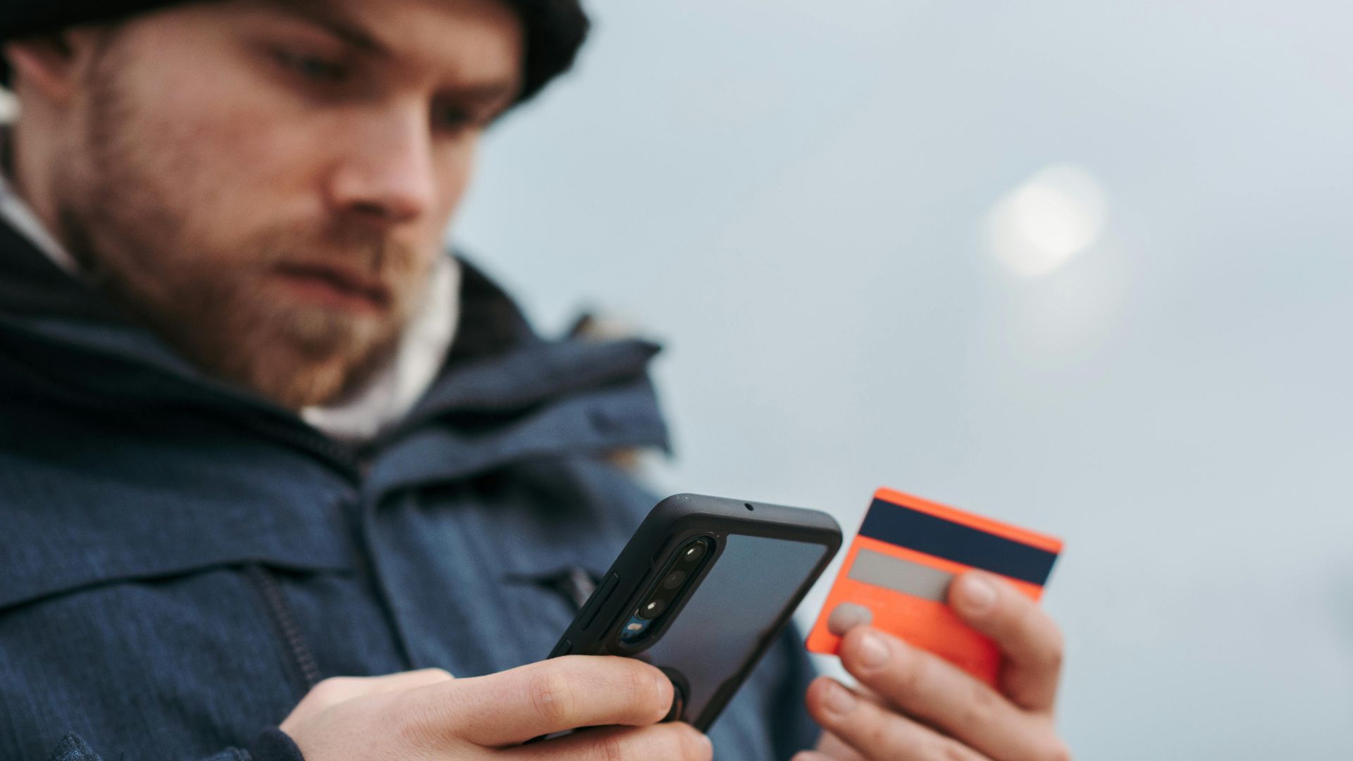 Crop male in outwear entering details of credit card on mobile phone while making online payment for purchase in daytime