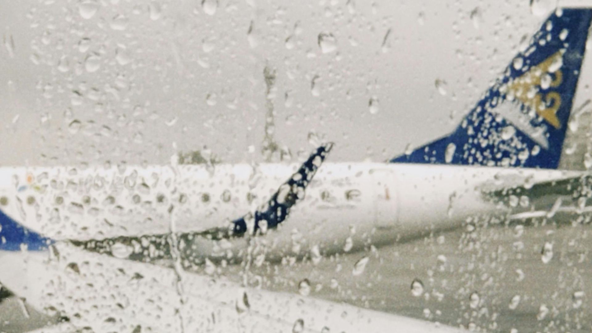 A rainy day view from an airplane window showing the wing and airport surroundings.