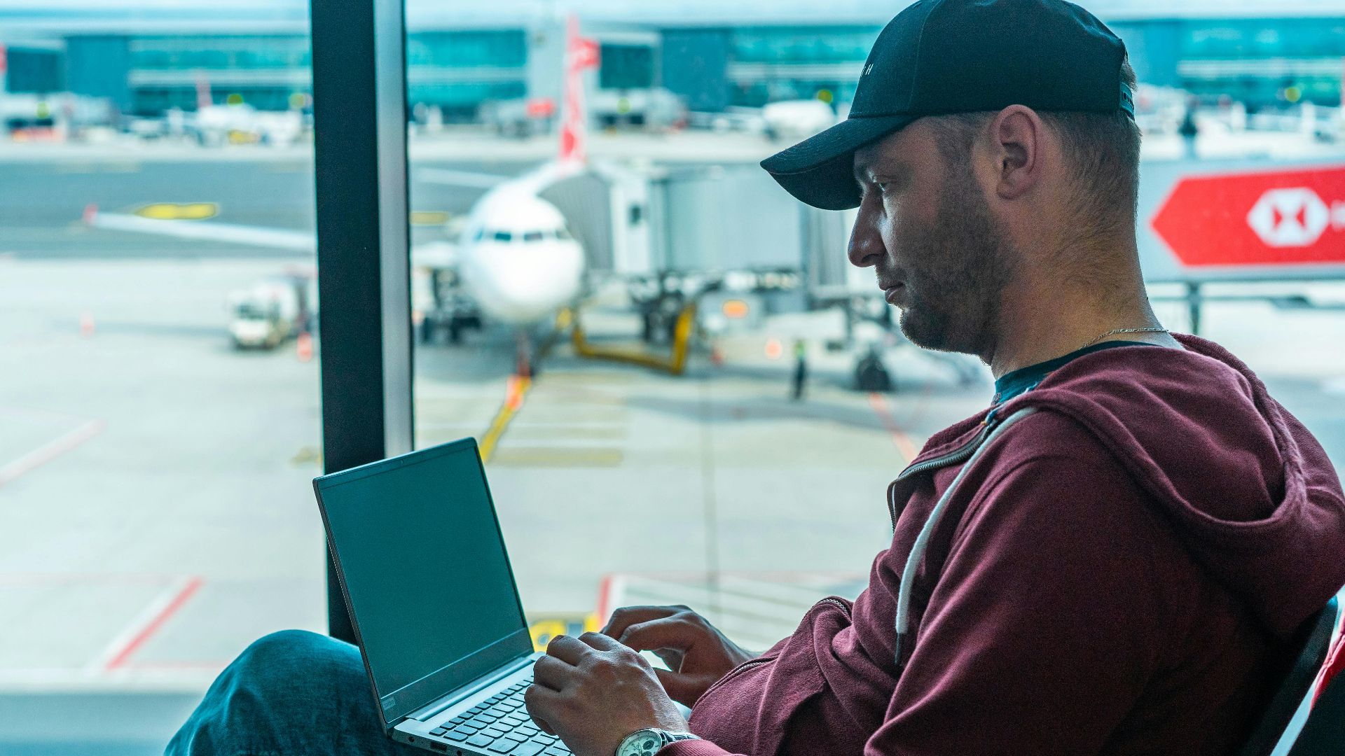 A man working on his laptop in an airport terminal, with a plane visible outside.