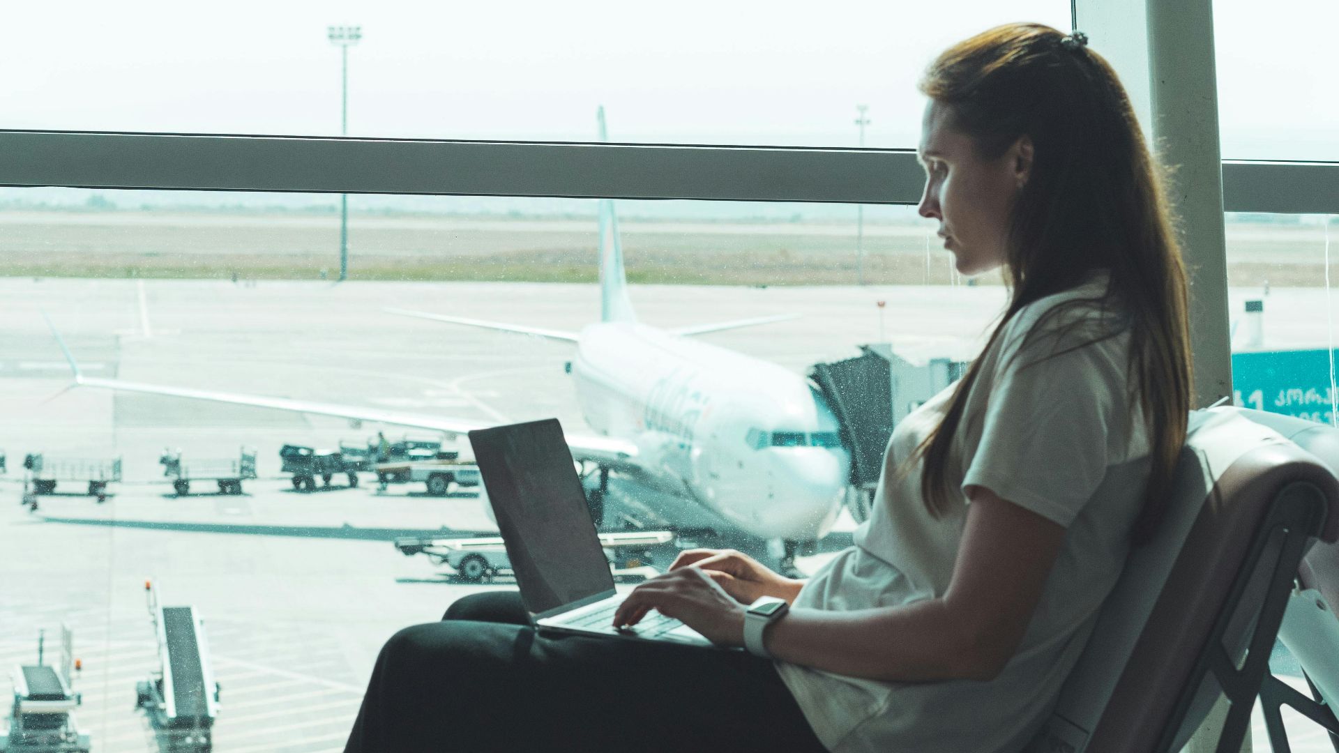 Side view of woman using laptop at airport, airplane visible outside through large glass windows.