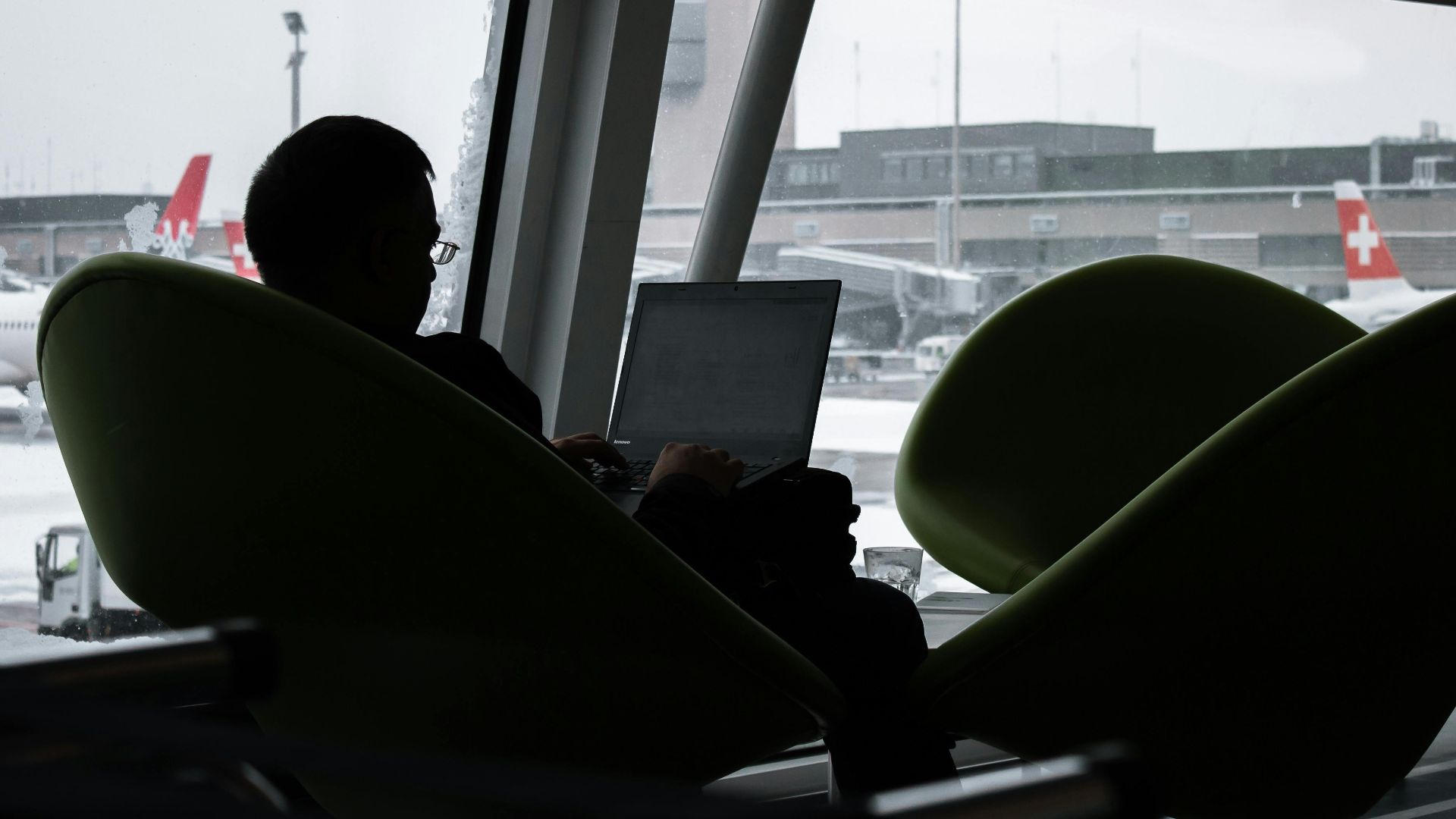 Silhouette of a man working on laptop at airport lounge with planes outside.