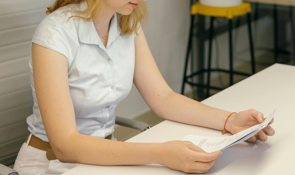 Woman in White Button Up Shirt Reading a Piece of White Paper while Sitting on a Chair