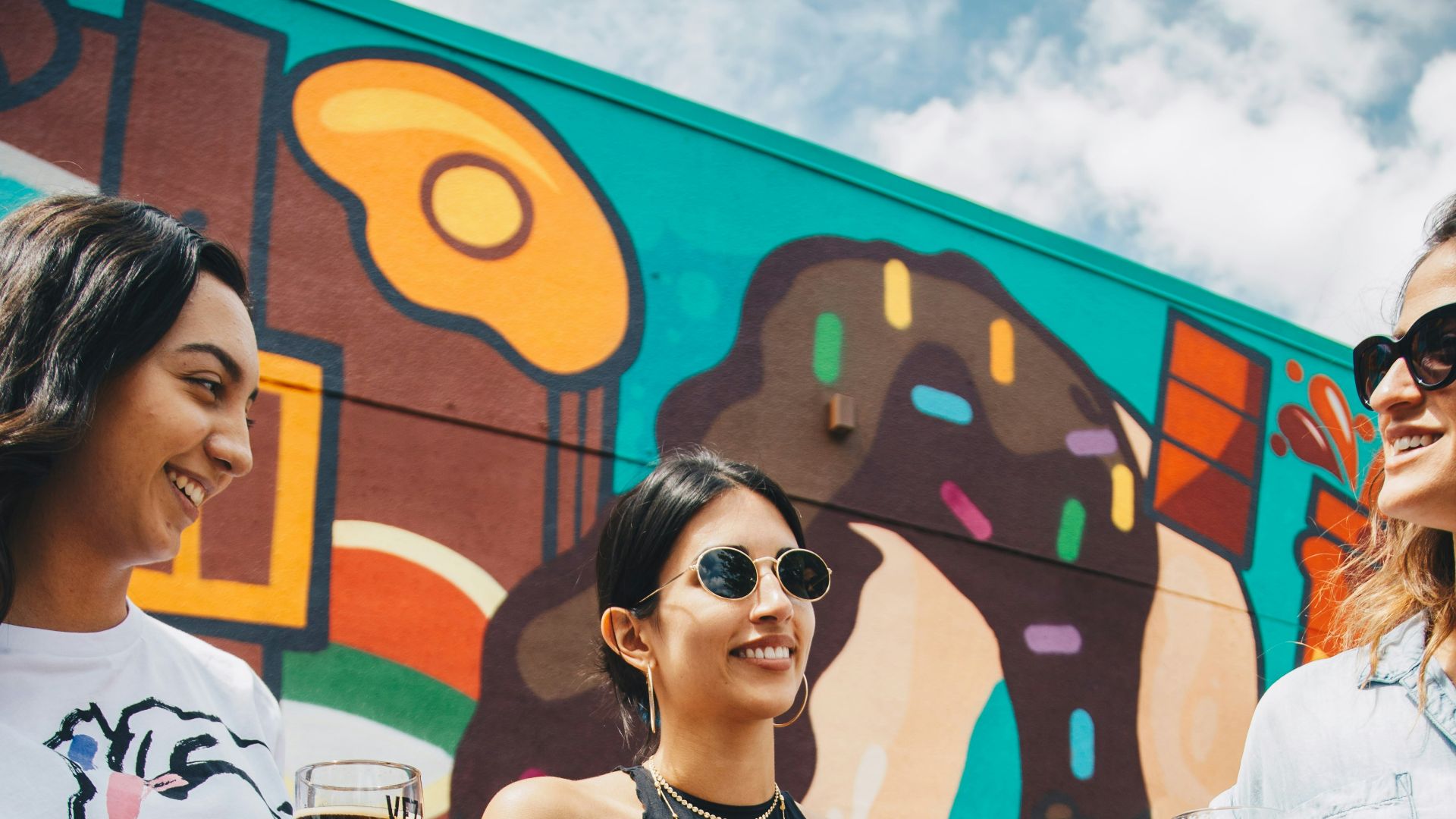 three women having a drink standing in front of wall graffiti