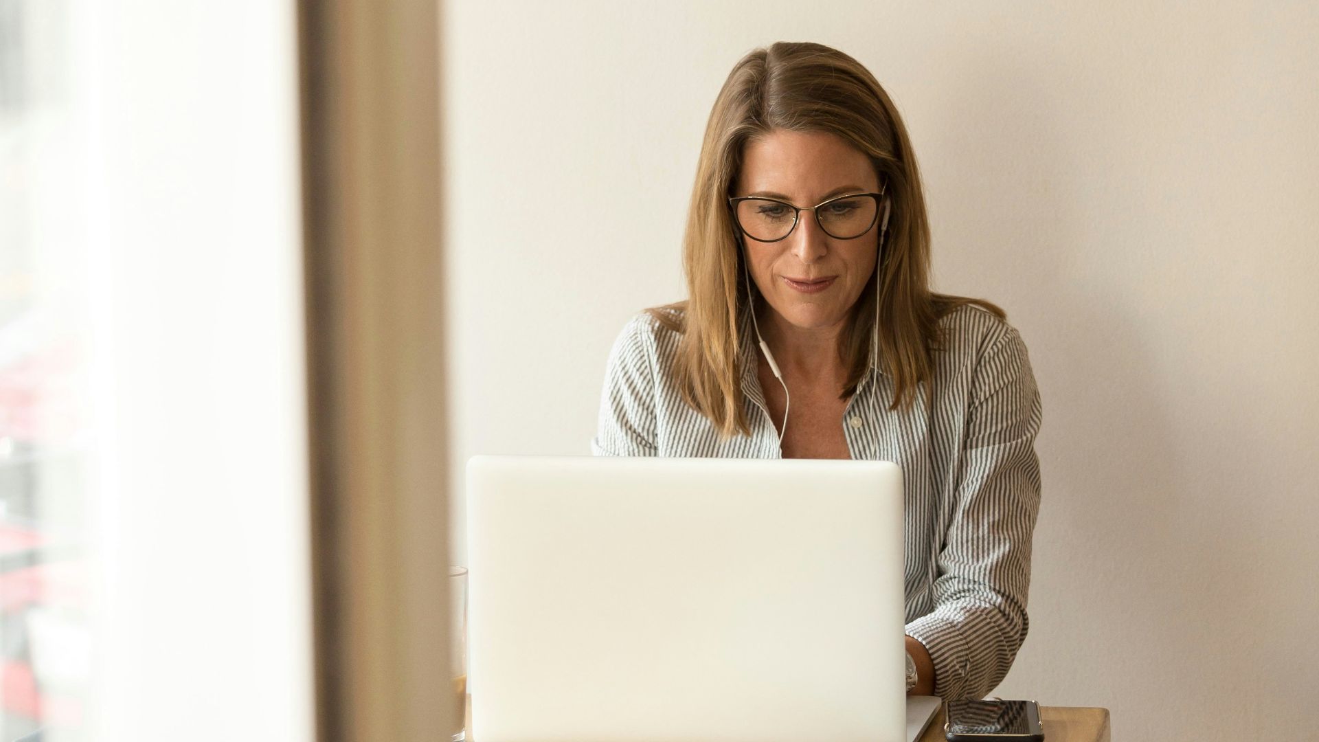 woman wearing grey striped dress shirt sitting down near brown wooden table in front of white laptop computer