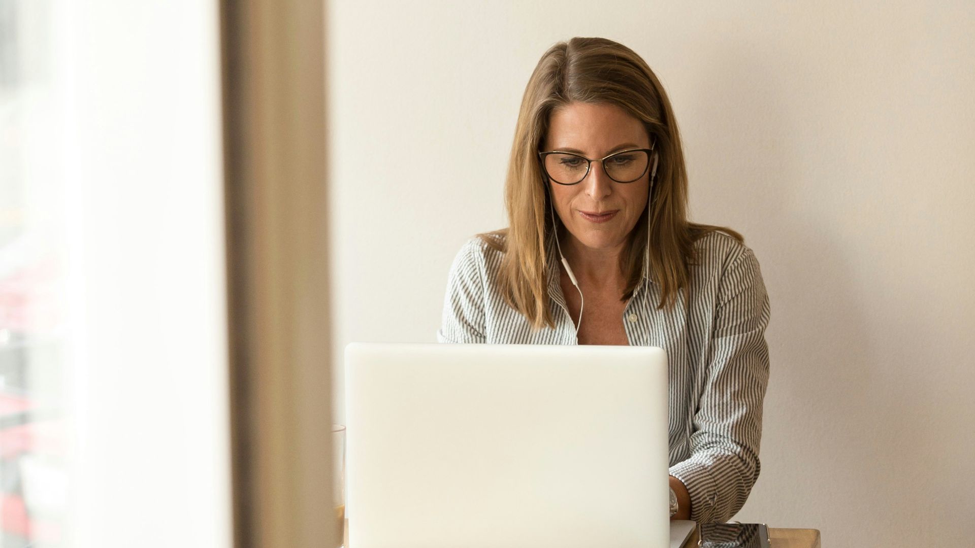 woman wearing grey striped dress shirt sitting down near brown wooden table in front of white laptop computer