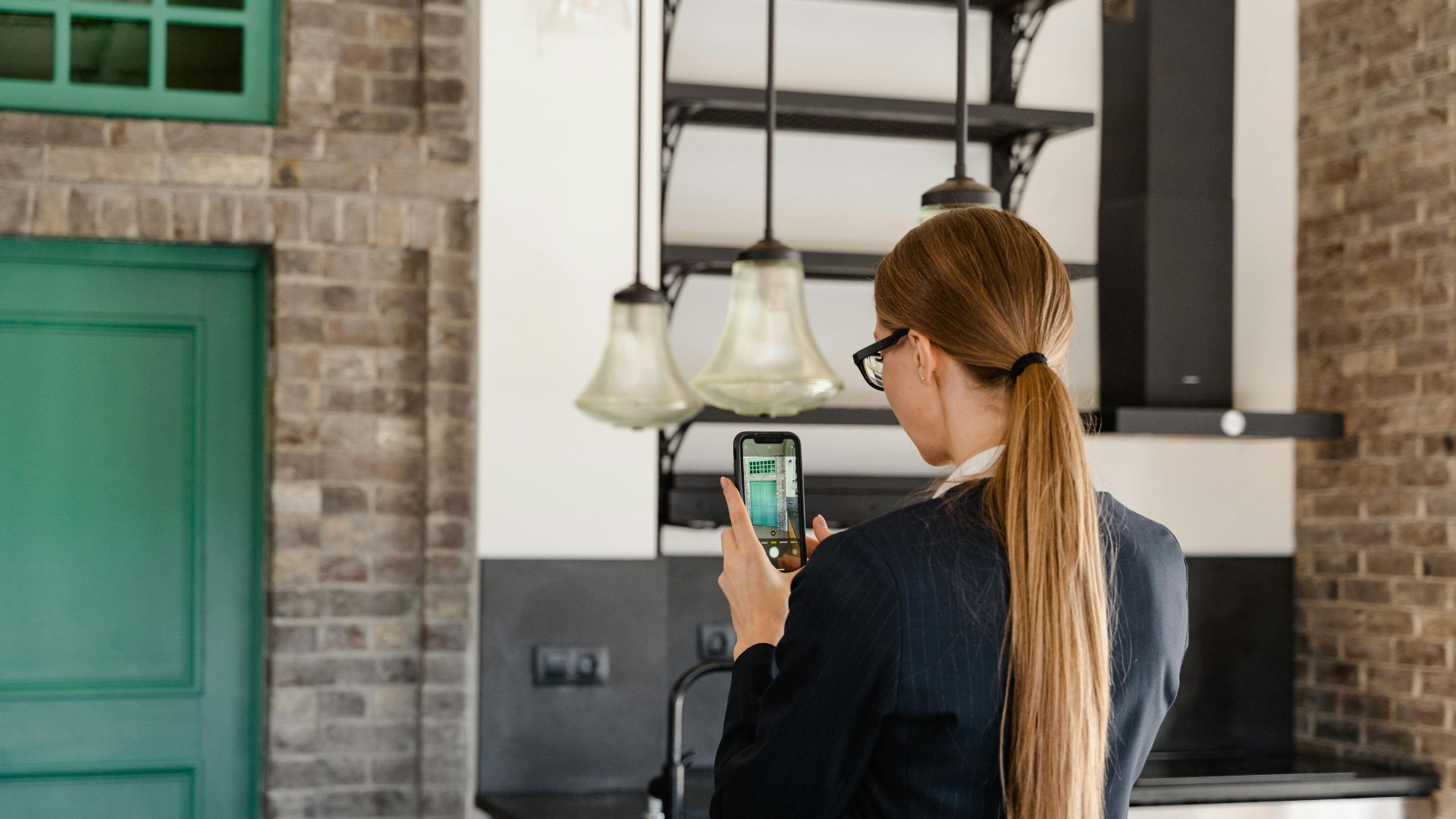 Rear view of a woman with long hair taking a photo with a smartphone in a stylish kitchen.