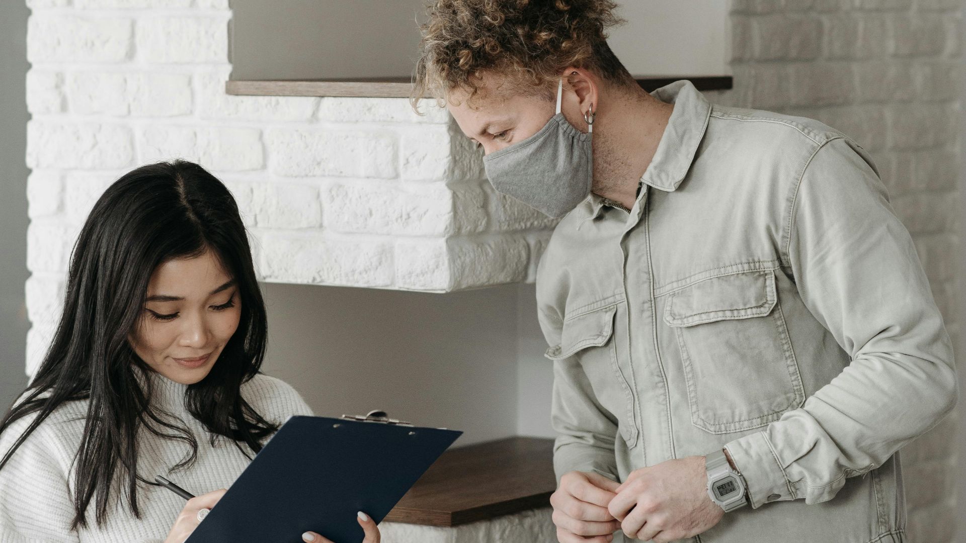 An Asian woman signs a clipboard for a delivery man wearing a face mask indoors.