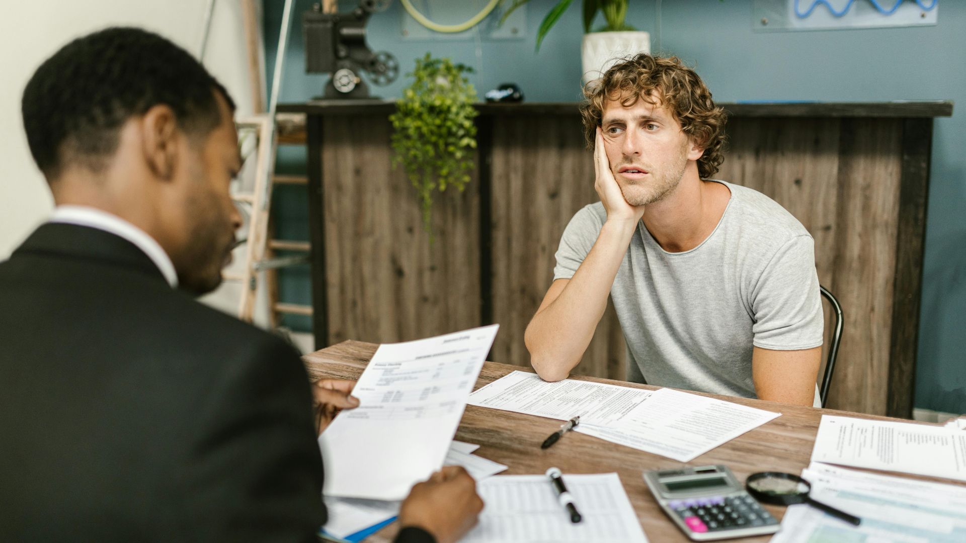 Man in office, appearing stressed during paperwork discussion, sitting opposite professional.