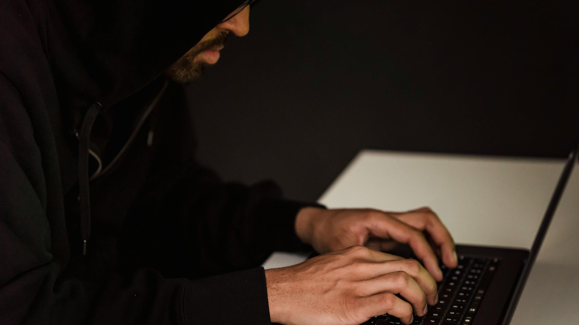 Crop bearded male hacker in black hood typing on netbook keyboard and browsing dark net in obscure room