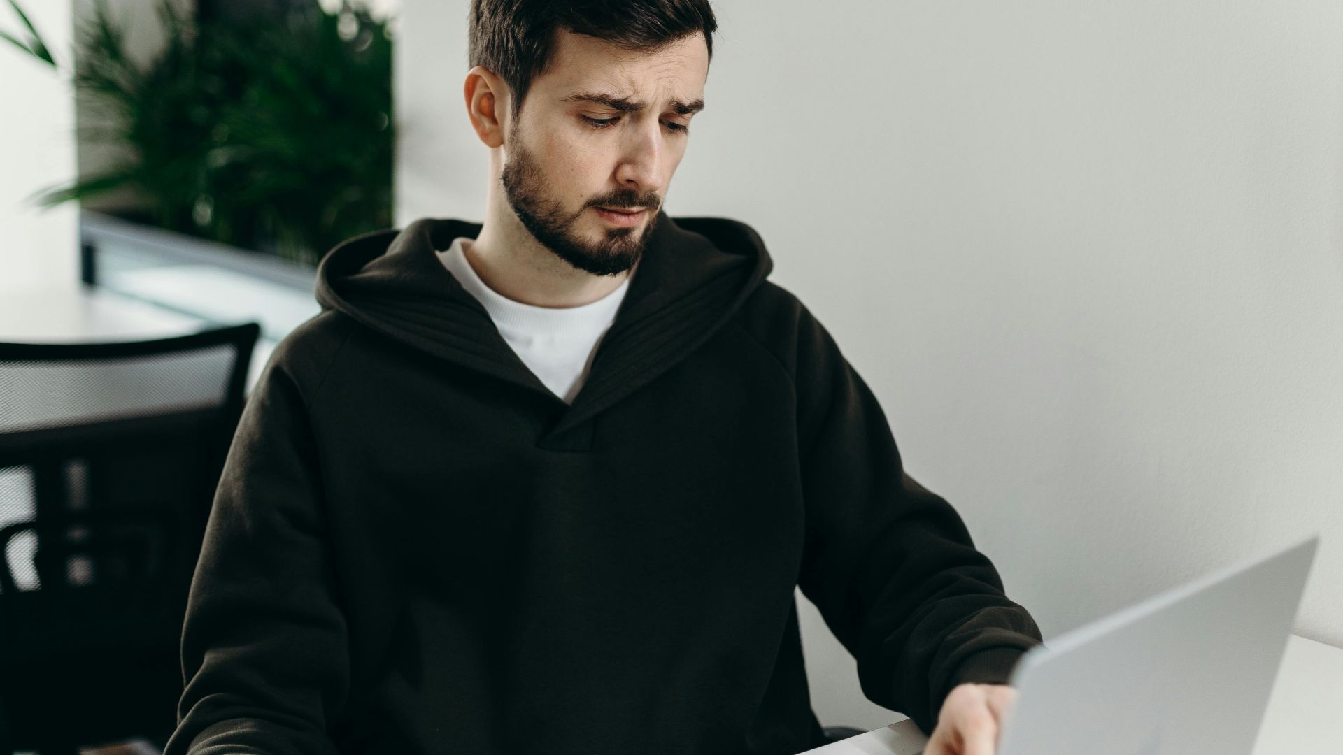 A young man in a hoodie working on a laptop at a home office desk, focused and thoughtful.