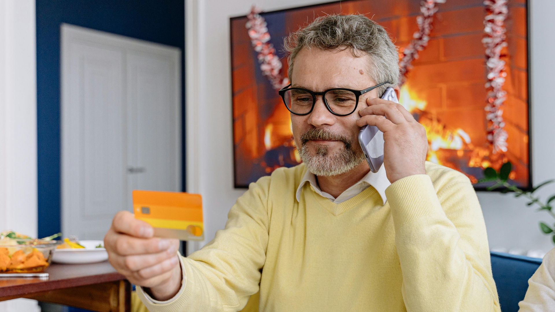 Elderly man using phone while holding a credit card indoors near a cozy fireplace.