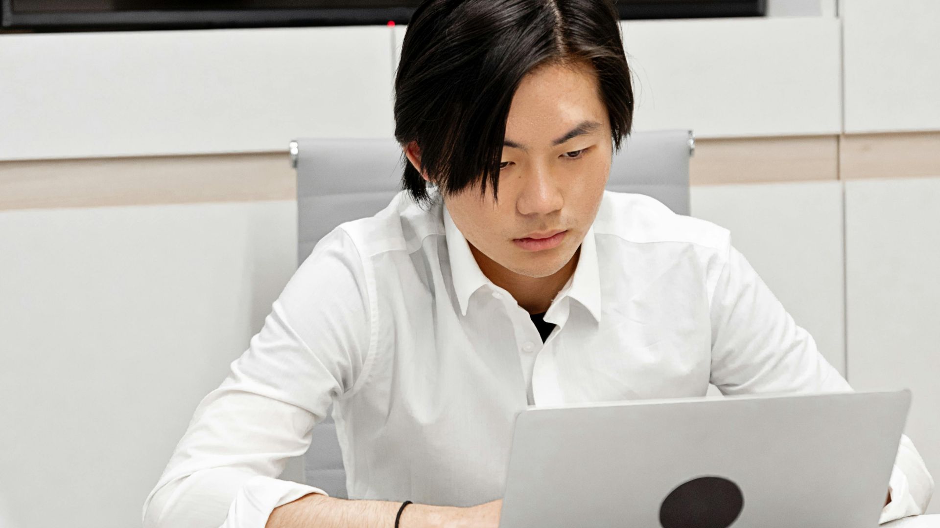 Asian man in white shirt working on laptop in modern office setting, focused and engaged.