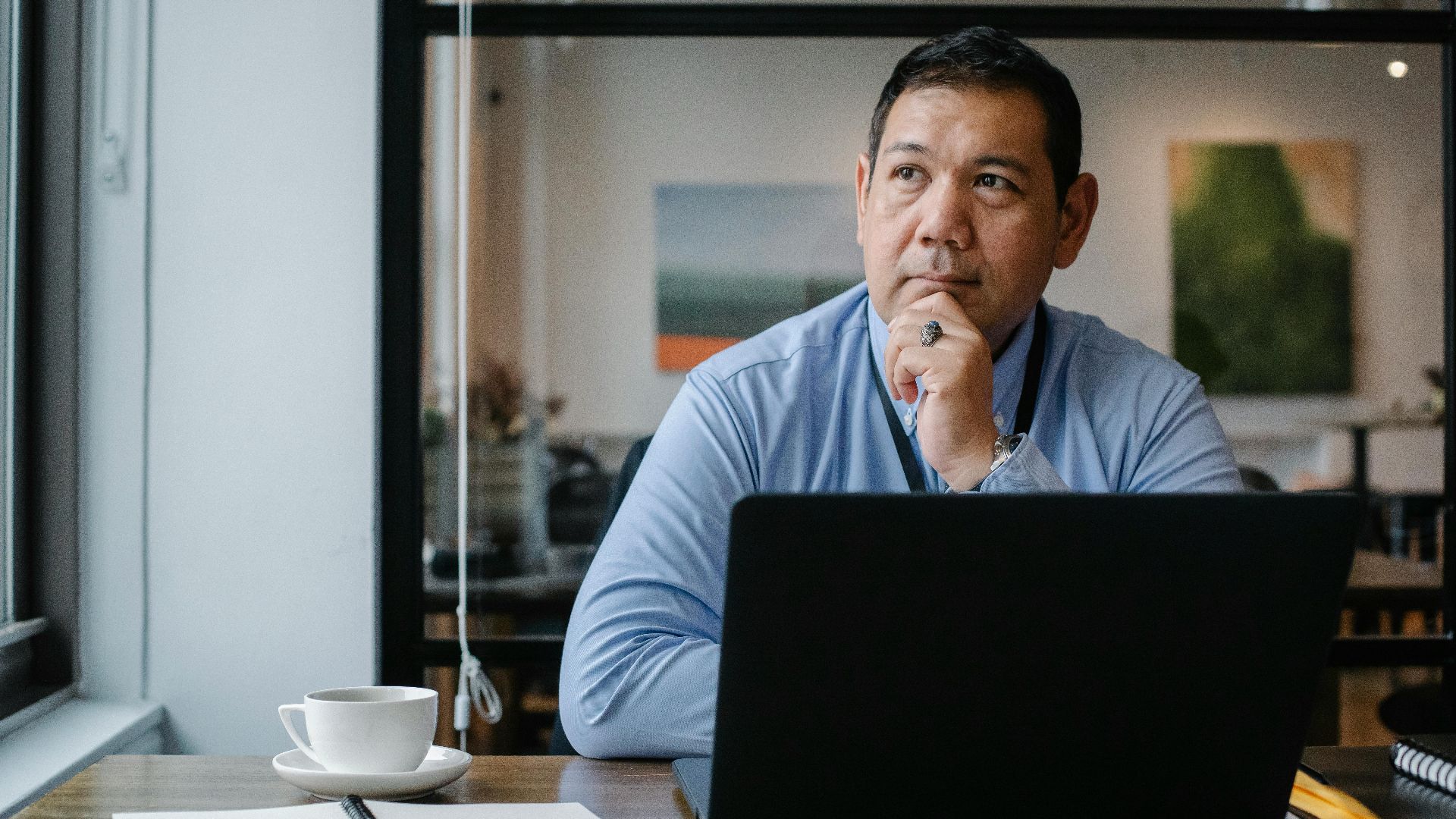 Concentrated ethnic adult male entrepreneur thinking on project while working at table with netbook and cup of drink