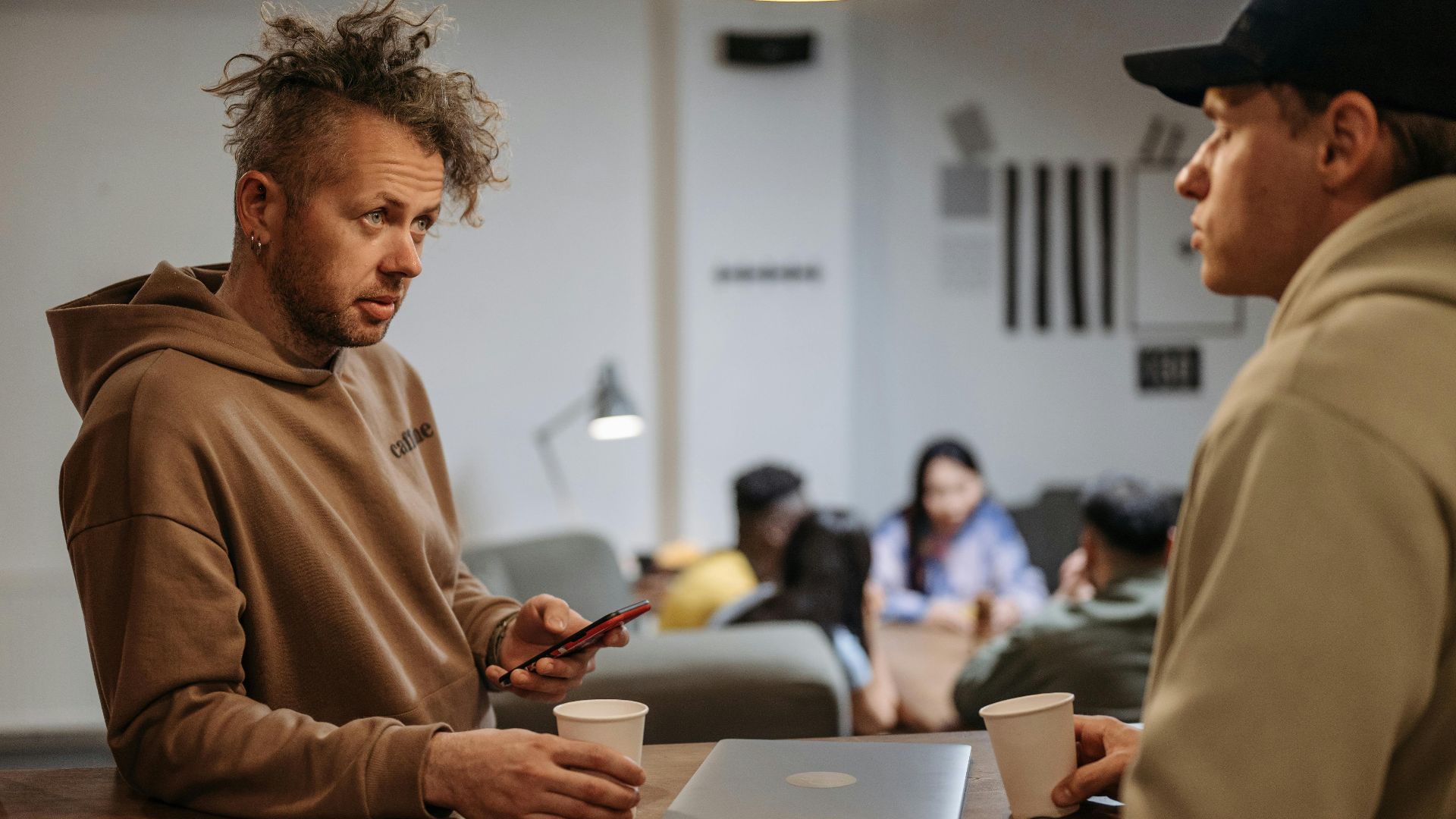 Two men in a casual office setting talking over coffee with laptops.