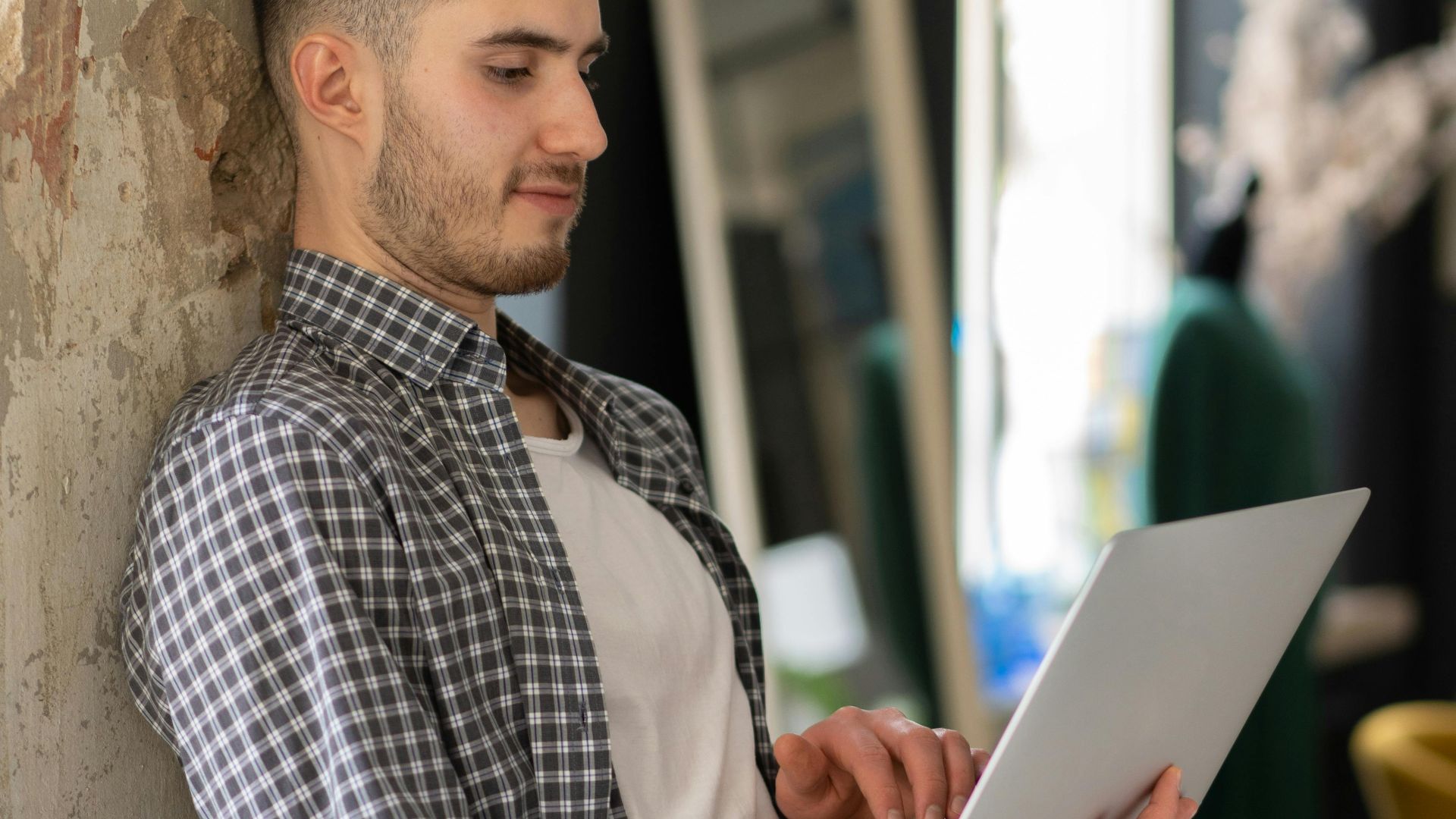 Stylish young man leaning on wall while working on a laptop at home.