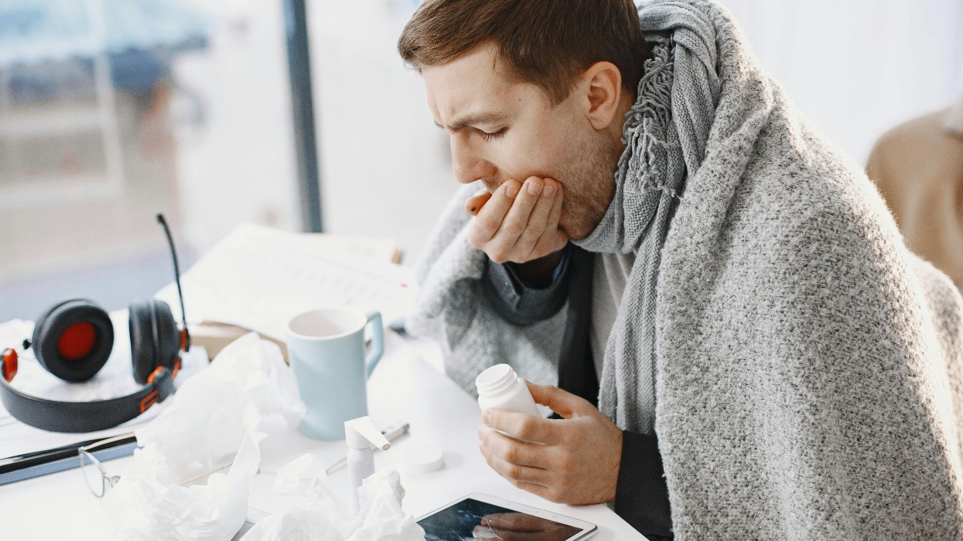 Man wrapped in blanket feeling unwell at home, surrounded by tissues and medication.