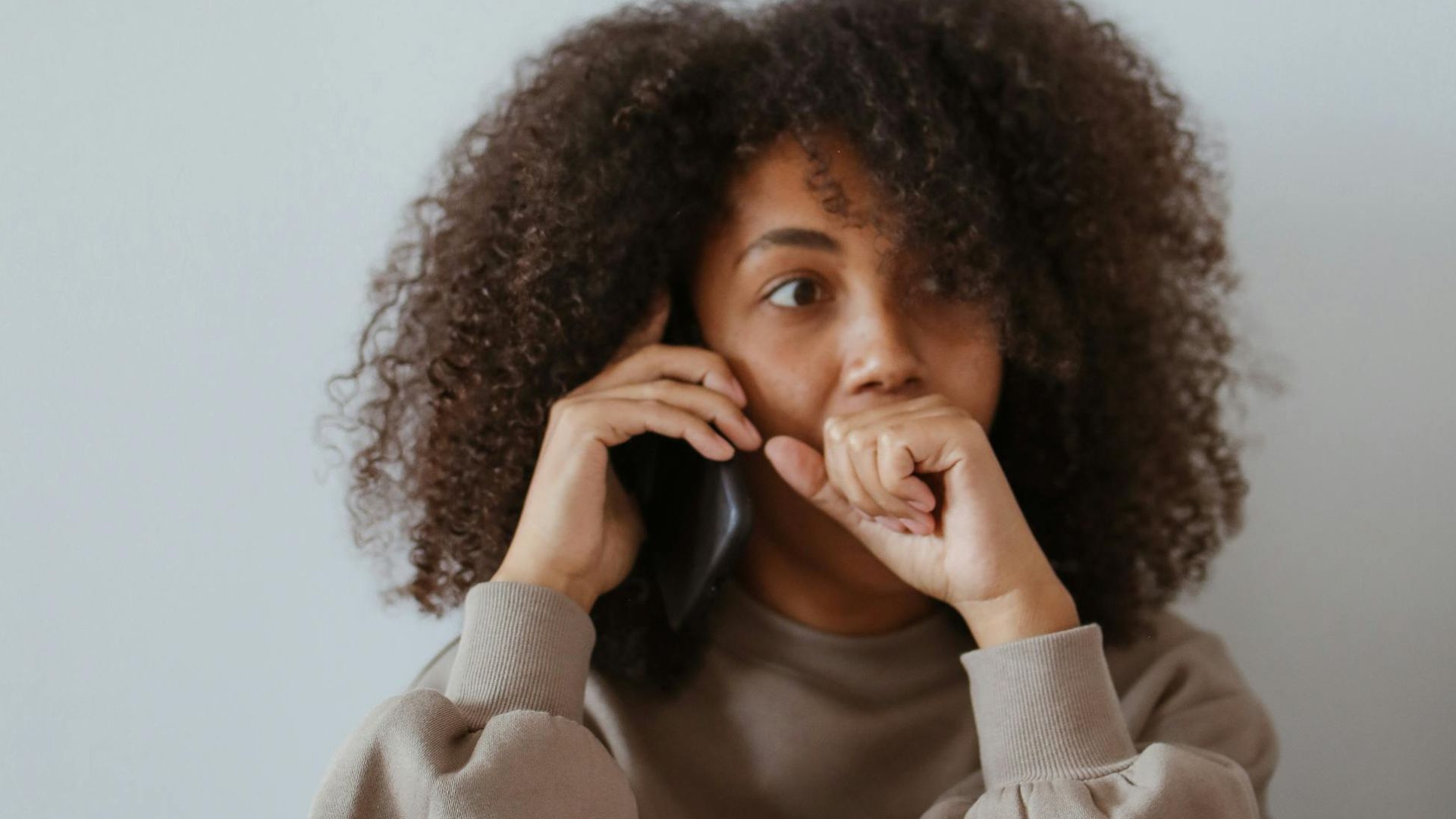A woman with afro hair in a brown sweater pondering during a phone call with documents around her.