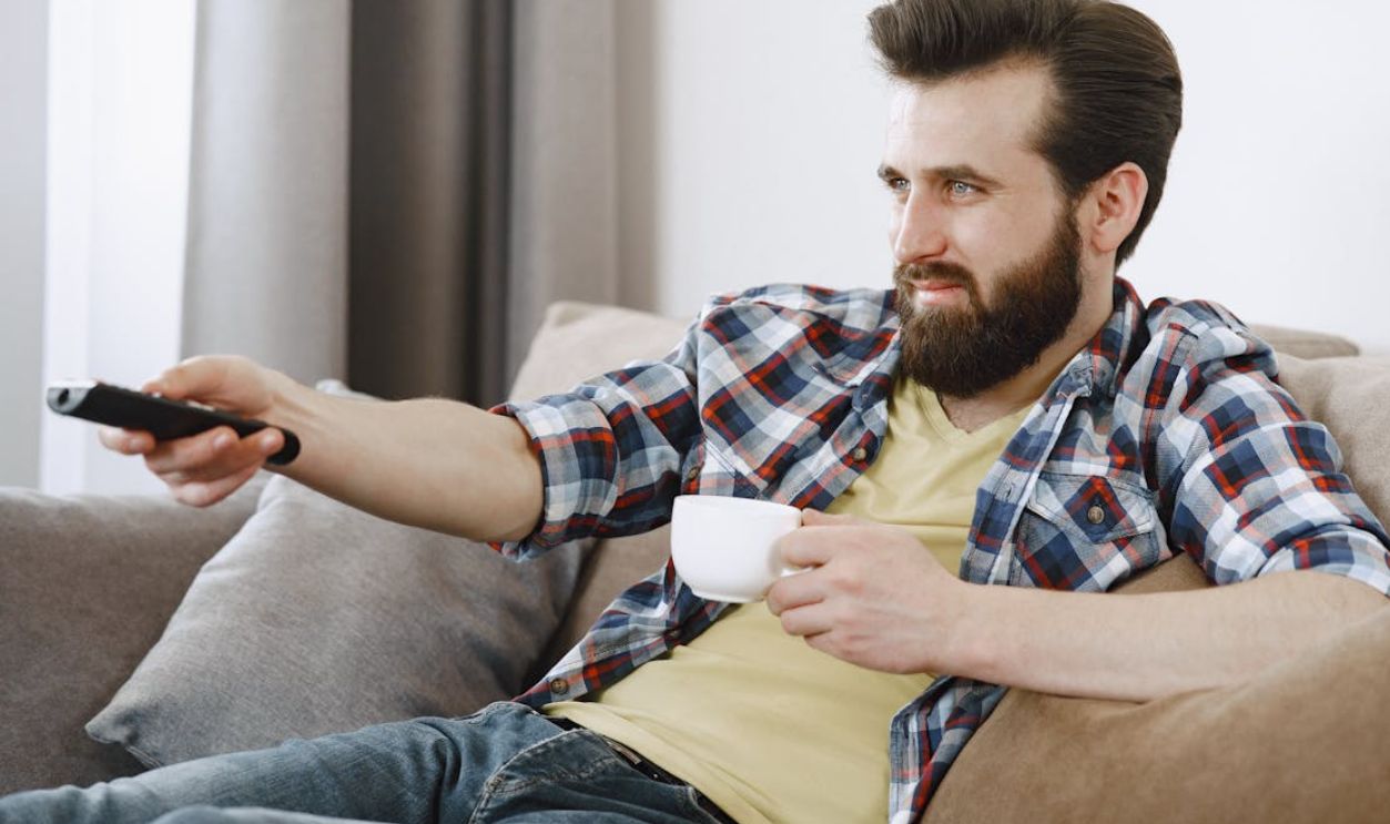 Caucasian man with a beard relaxing at home, holding a cup and watching TV on a comfortable sofa