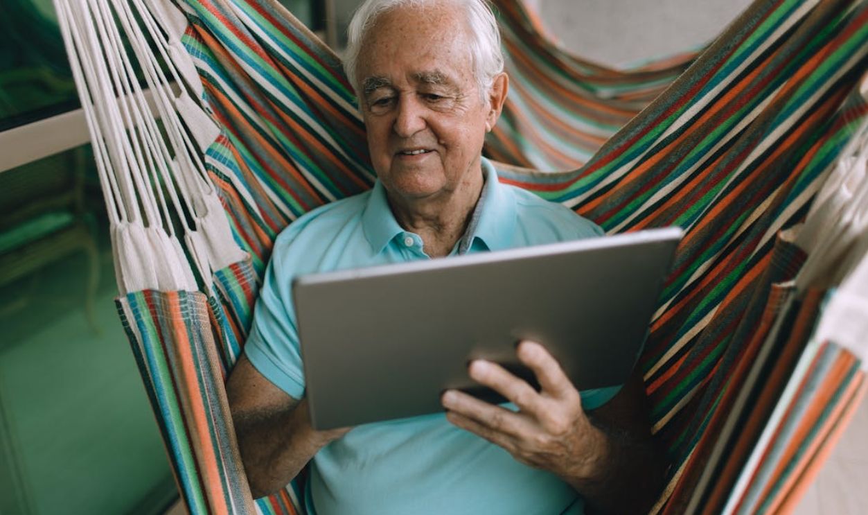 Elderly man enjoying leisure time in a hammock while using a tablet indoors.