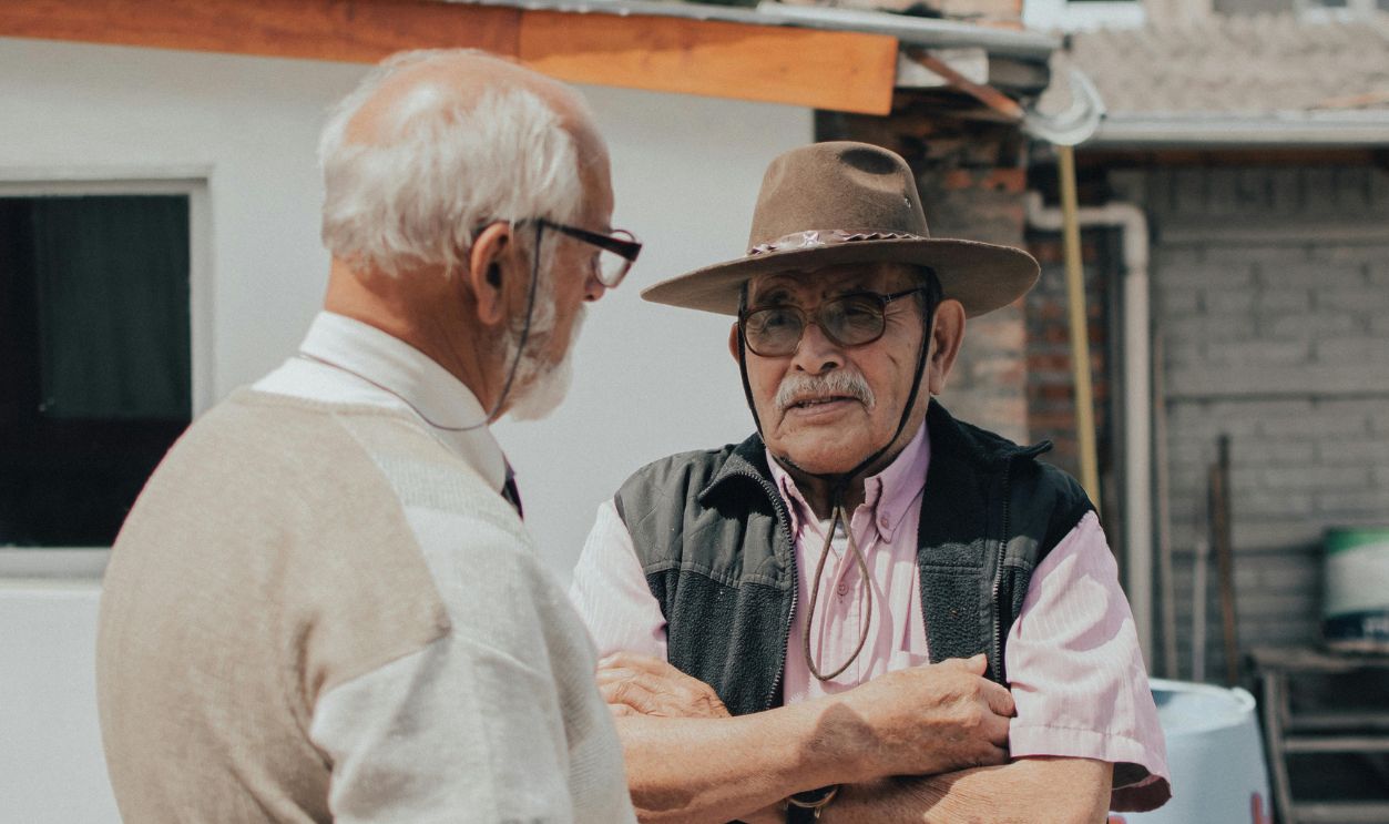Two senior men engaged in conversation outside a rustic building on a sunny day.