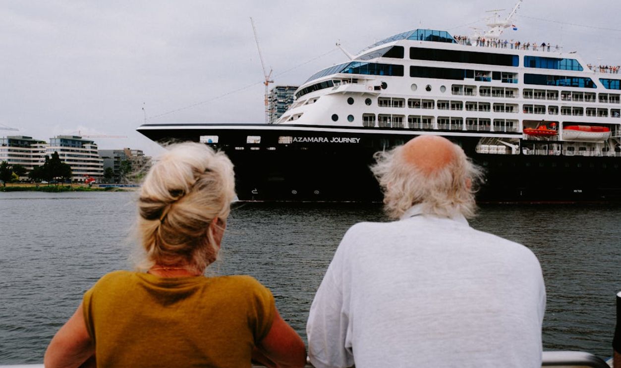 An elderly couple watches a large cruise ship in a bustling harbor.