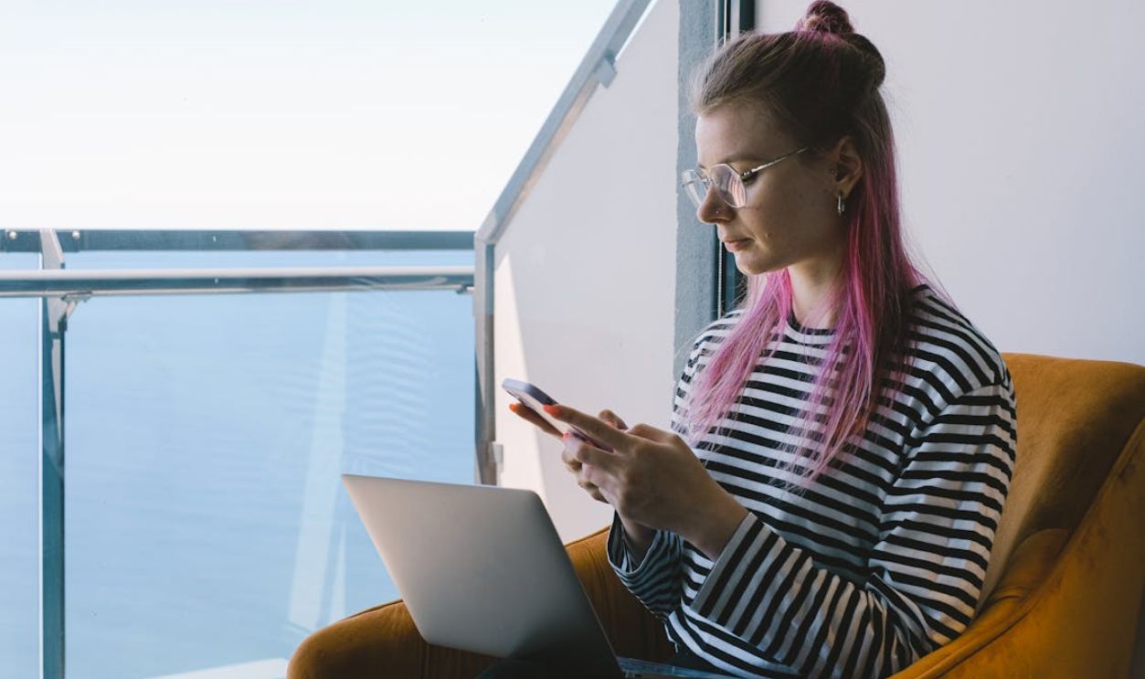Young woman working remotely, using laptop and phone, enjoys sea view.