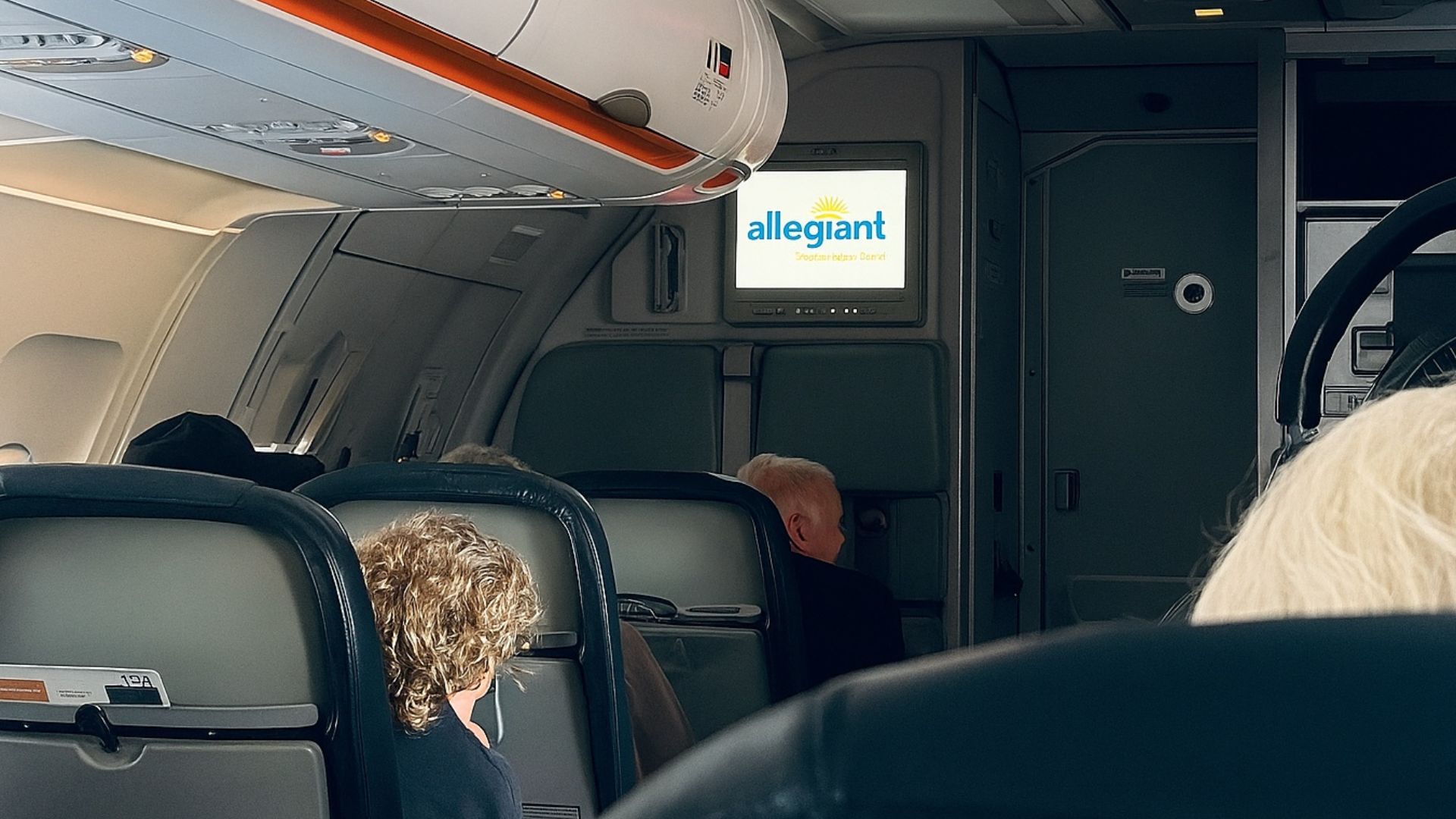 Interior of an Allegiant Air aircraft cabin, viewed from the rear toward the front, showing seating layout, overhead compartments, and the Allegiant logo displayed on the cabin screen.