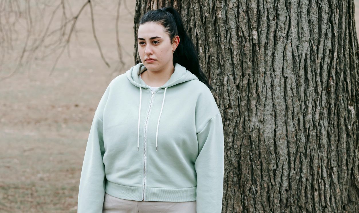 Young woman in a hoodie stands thoughtfully against a tree in a leafless park setting