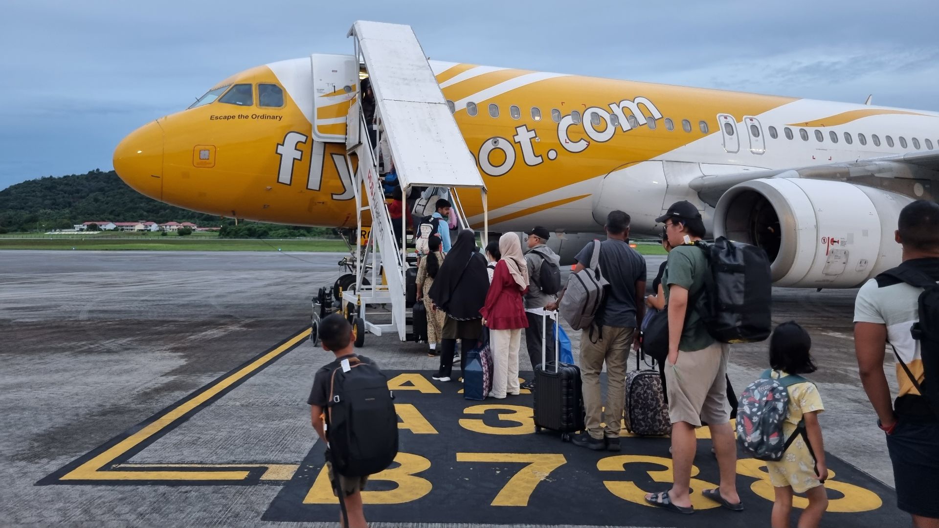 A photograph showing passengers on the apron of Langkawi International Airport queuing to board an Airbus A320 aircraft under Scoot Airlines. The flight is TR477 from Langkawi (LGK) to Singapore (SIN) and was delayed from its scheduled departure time of 1