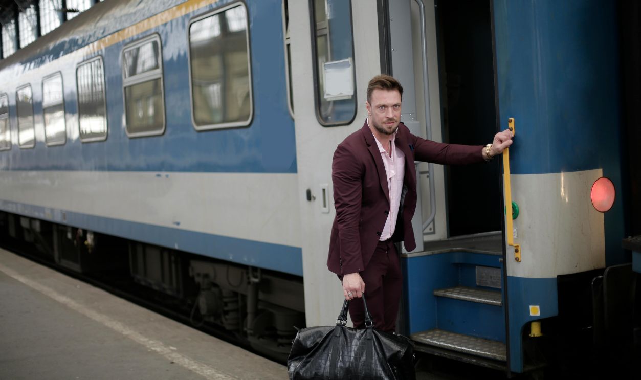 A well-dressed man in a burgundy suit boards a train holding his black travel bag