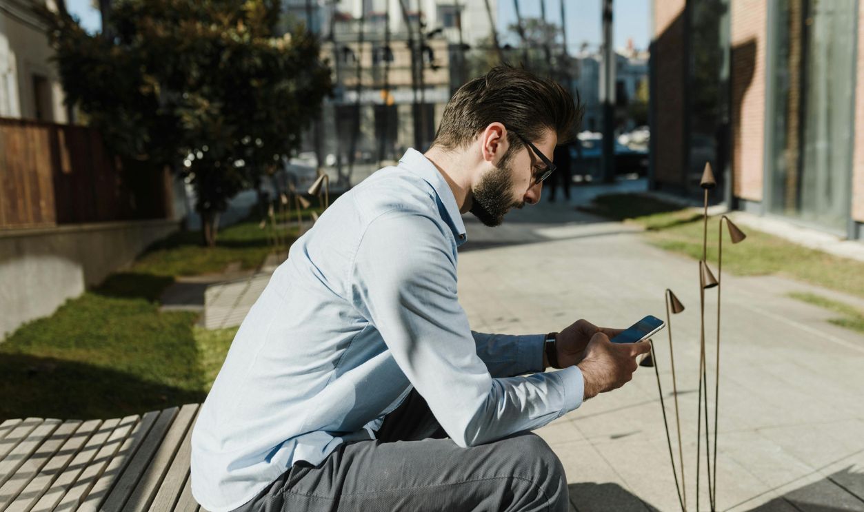 Adult man with beard sitting outside on a bench using smartphone. Side view
