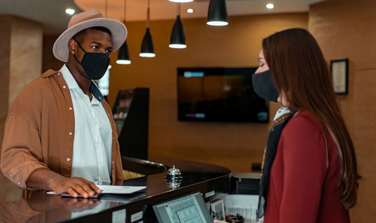 A man and woman in face masks checking in at a hotel reception, emphasizing safety protocols
