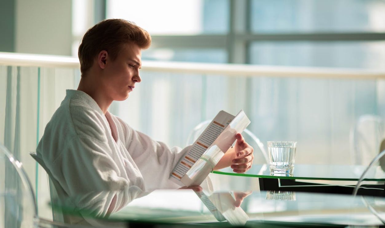 Man in bathrobe reading by a glass table indoors, reflecting tranquility.