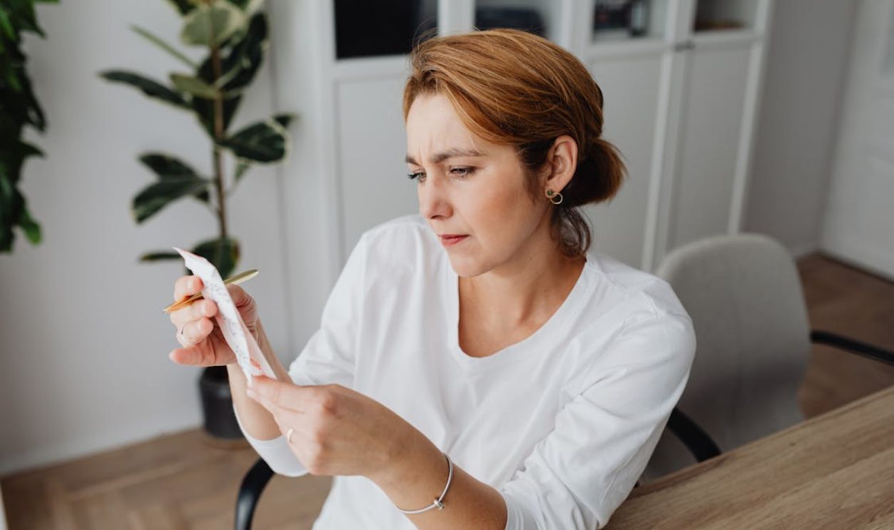 A concerned adult woman examines a receipt while sitting at her office desk, indicating financial scrutiny.
