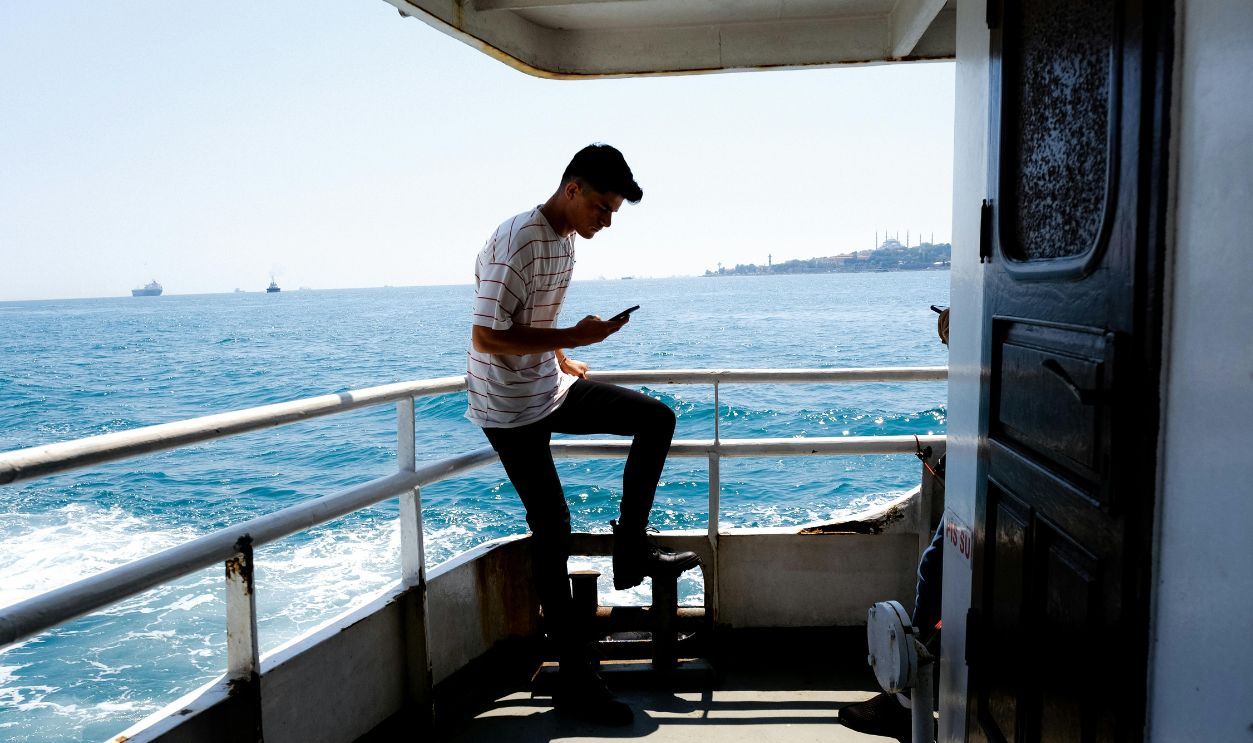 A man stands on a boat, looking at his phone with a scenic sea backdrop on a sunny day.