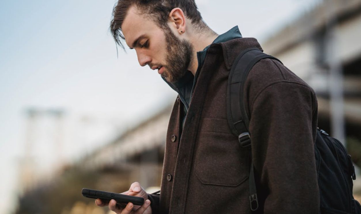 A young man with a backpack intently looks at his smartphone in an urban setting