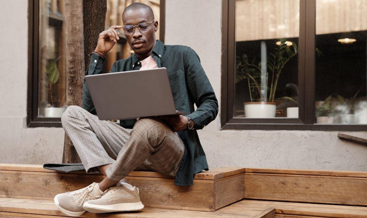 Stylish man with glasses working on a laptop while sitting on wooden steps outdoors