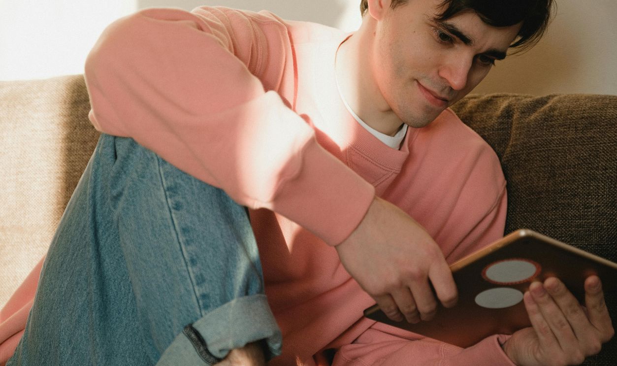 A man in a pink sweater sitting on a sofa, using a digital tablet