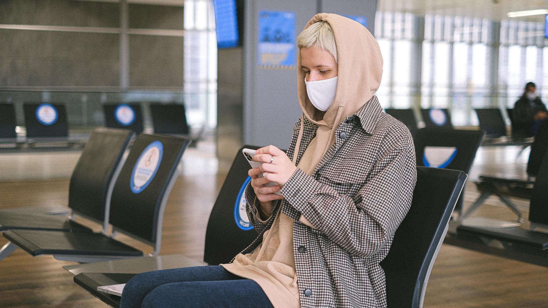 A woman wearing a face mask and a hood sits in an airport waiting area, using her smartphone.