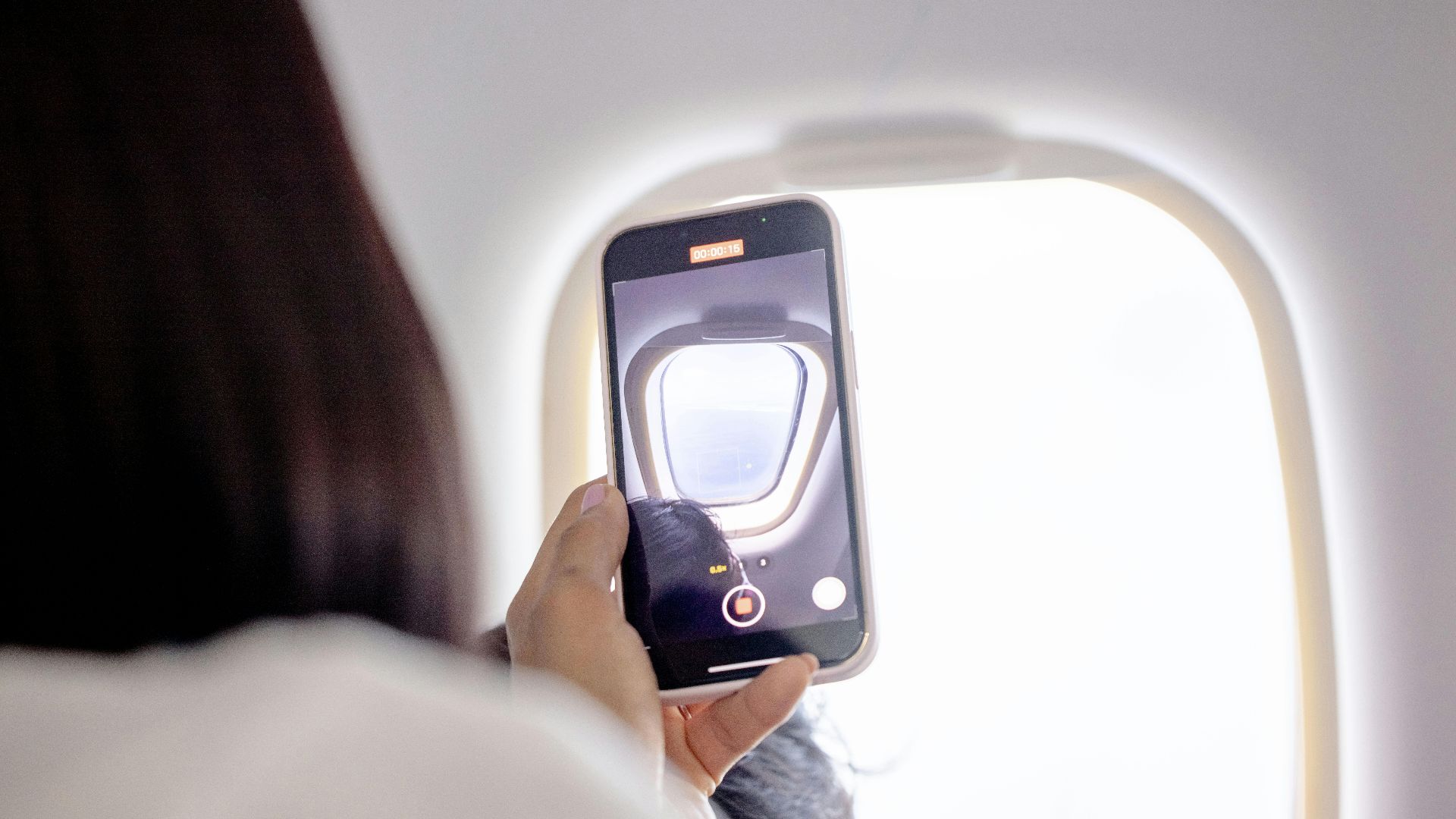 A woman records a video of the sky through an airplane window using her smartphone.