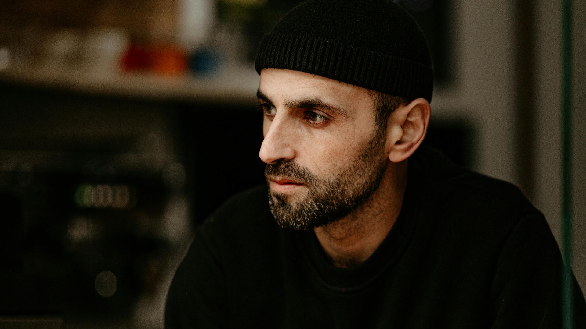 A contemplative man in a black beanie sitting at a wooden café table, looking away pensively.