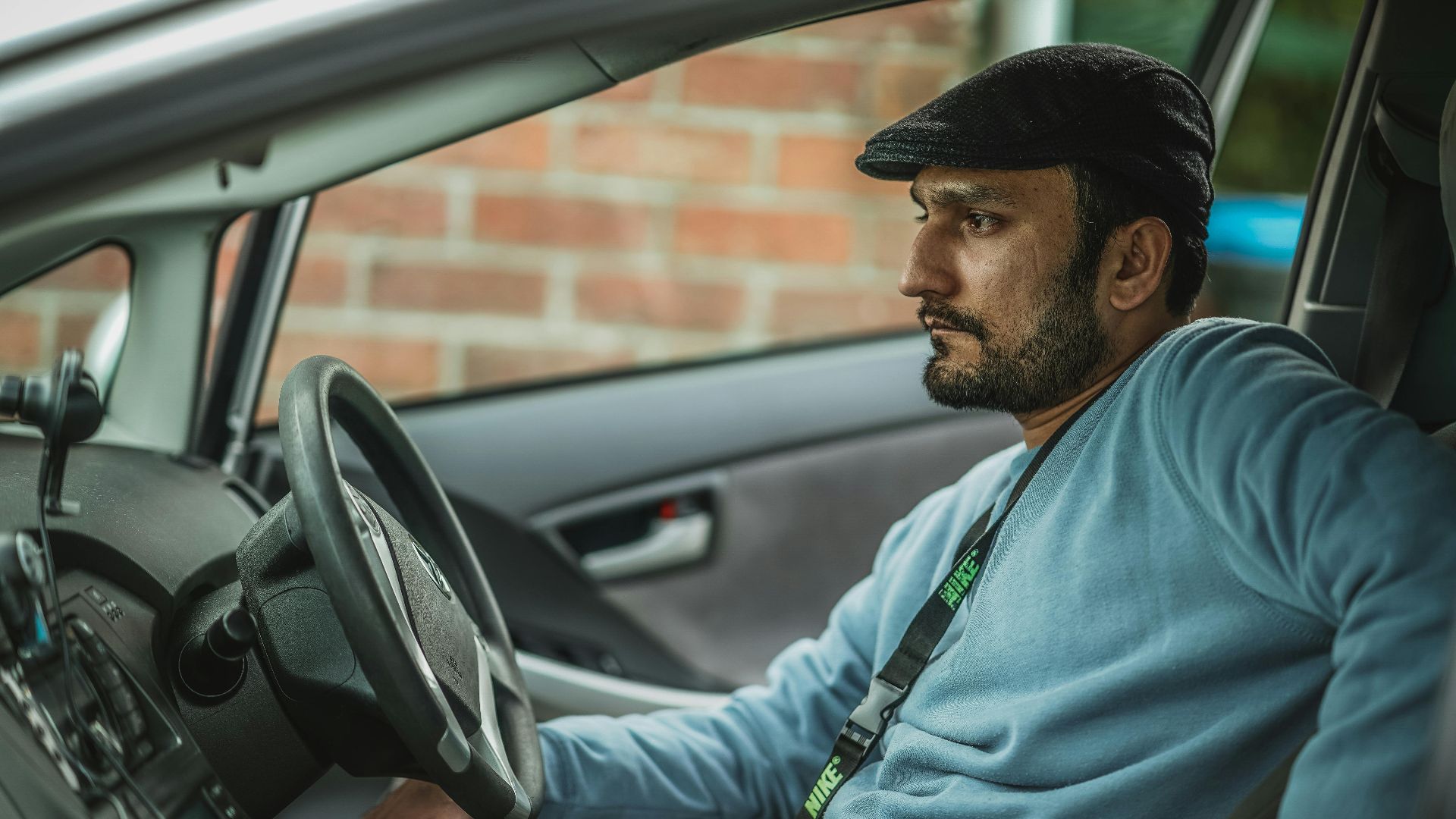 A bearded man wearing a hat and blue shirt sits thoughtfully behind the wheel of a car.
