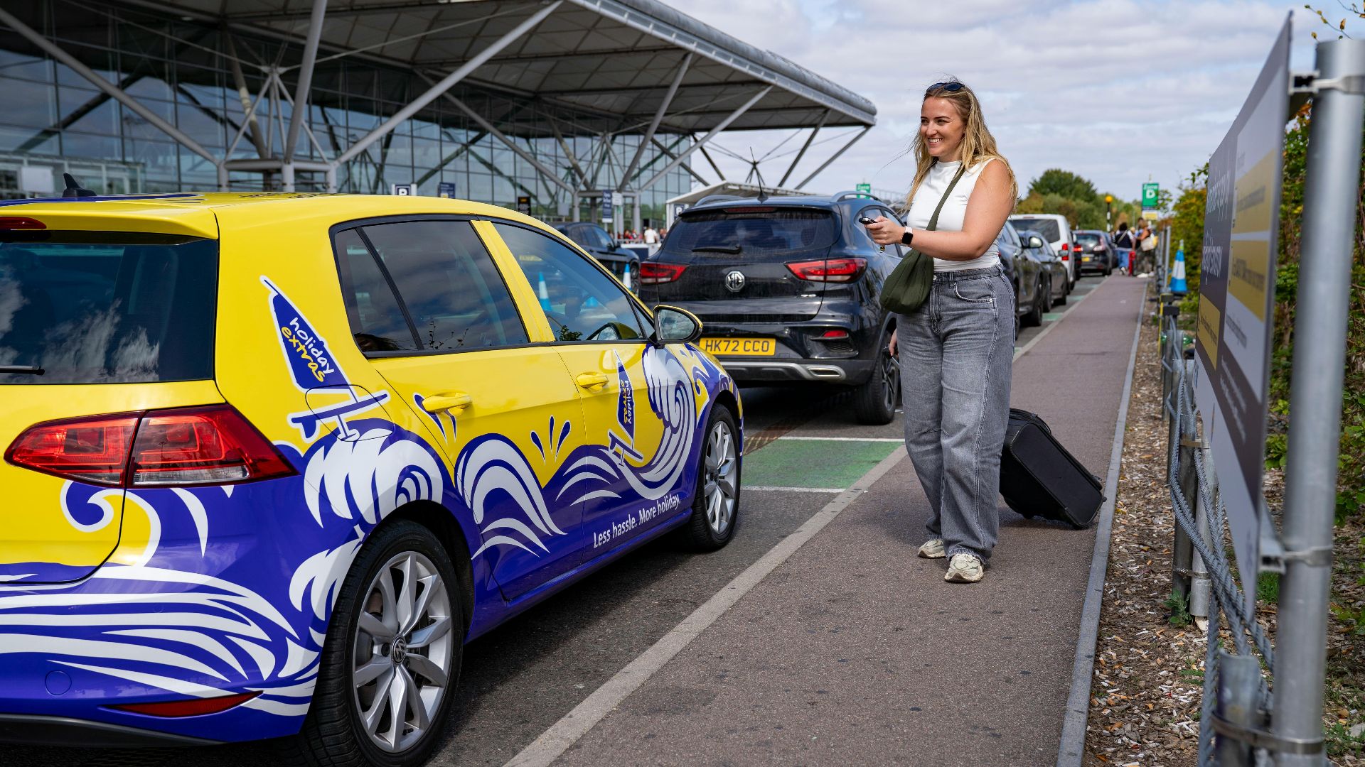 Woman smiling by a colorful car at Stansted Airport's valet parking area.