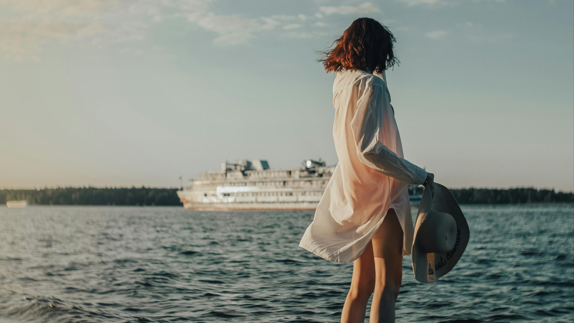 Woman in white shirt stands on shore watching a cruise ship on a sunny day.