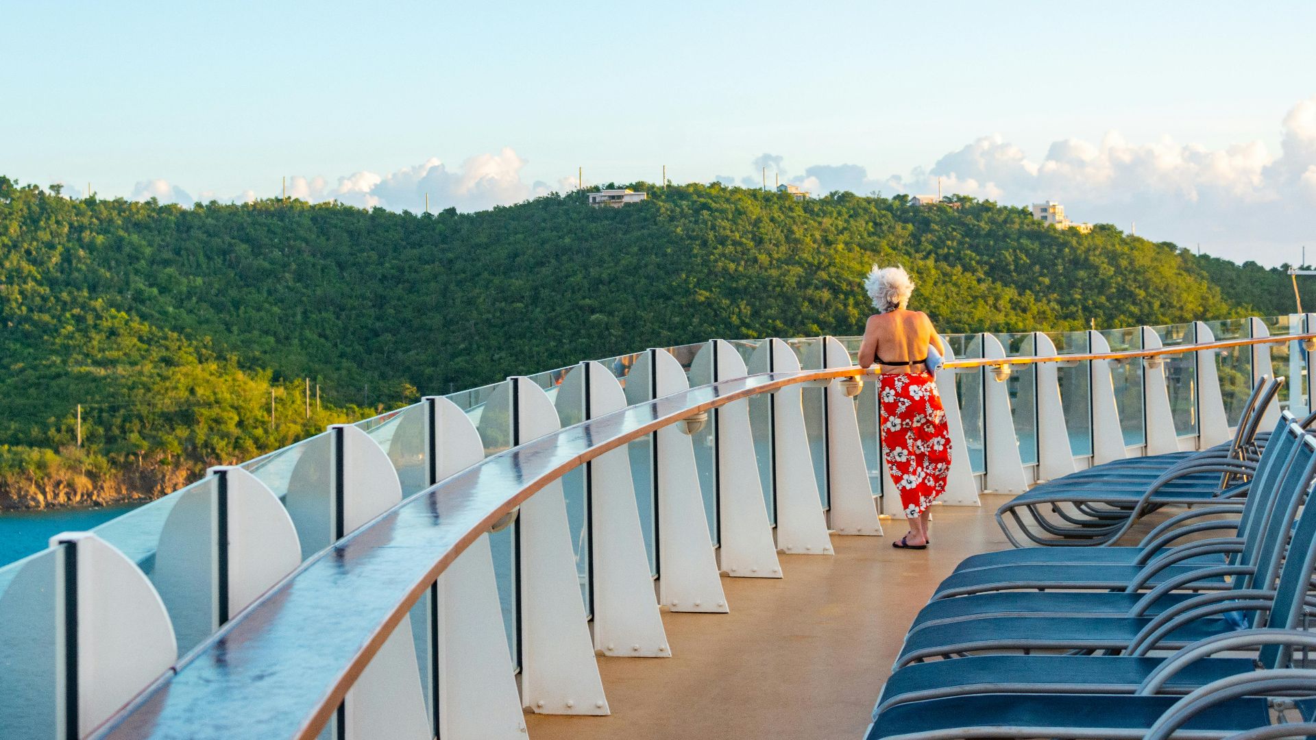 Senior woman enjoying a serene view from a cruise ship deck, surrounded by lush hills and ocean.