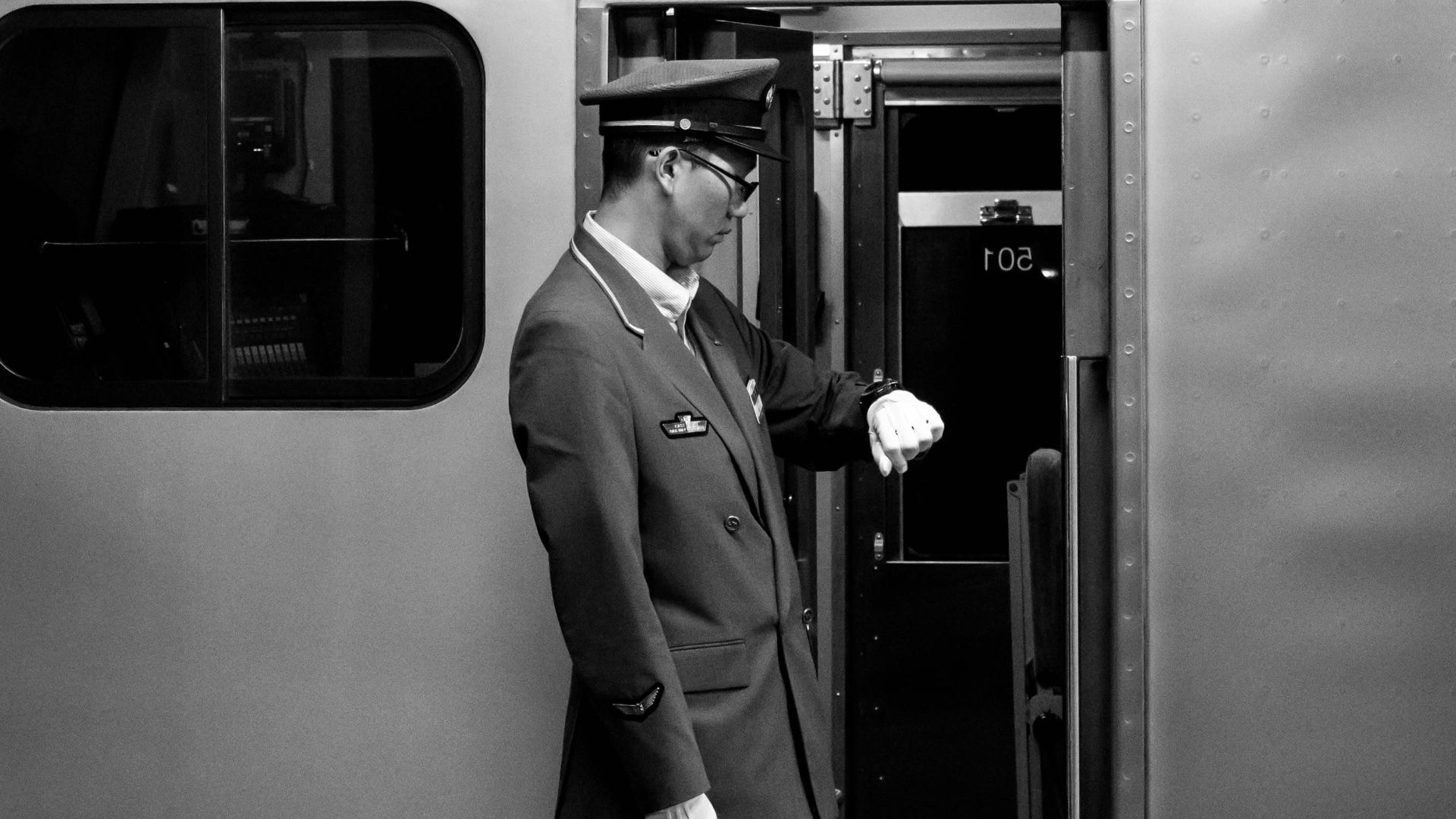 Black and white side view full body of train driver in cap and uniform looking at wristwatch while waiting for departure at railway station