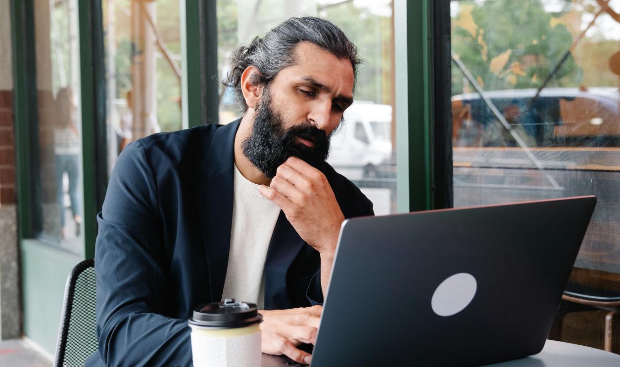 A focused man with a beard works on his laptop at an outdoor cafe