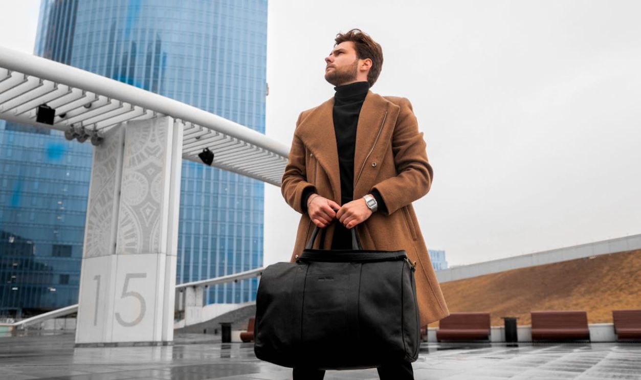 Fashionable man in coat holding a duffel bag with modern architecture in background