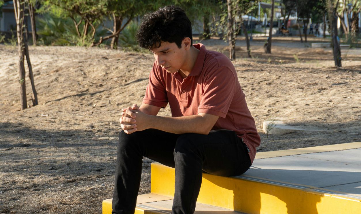 A young man sits on yellow steps in a park in Piura, Peru, lost in thought during a sunny day